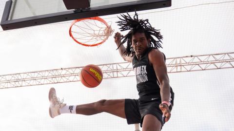 A basketball player leaps into the air and throws the ball into the hoop. One of his legs is raised in the air, and his hair flies up around his head. 