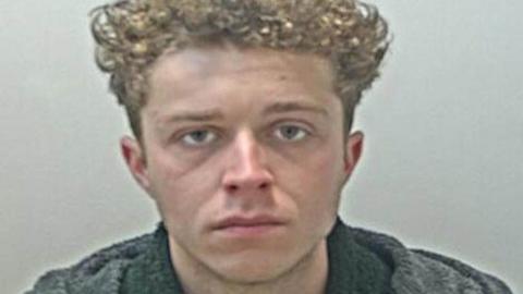 Police custody image of Frankie Bromley, who has light brown curly hair nd is wearing a black jumper and grey coat.