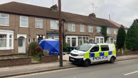 A police vehicle parked on the side of the road next to some terraced houses. A blue police tent is in front of a property's front door and the driveway is taped off with blue and white police tape.