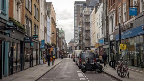 A street in Soho in central London. On both the left and right are a row of shops with people walking on the pavements outside. In the middle of the picture is a road with vehicles parked on the right.