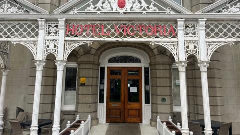 The entrance to the Hotel Victoria is seen with brown wooden doors, pillars and the signage above the door 