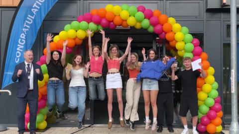 A row of eight students jumping into the air with their arms up in front of a multicoloured balloon arch. The headmaster is standing on the left celebrating. They are in front of a school building.