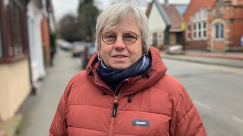 A woman with grey hair and round brown glasses is wearing a blue scarf and red-brown coat. She is standing against a slightly blurred background of a town street.