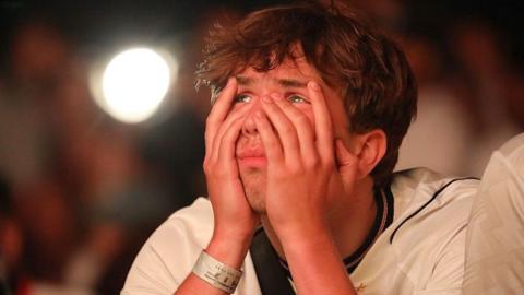A man wearing an England shirt partially covers his face with his hands and looks close to tears.