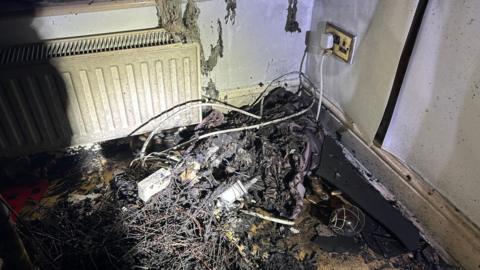 A bundle of wires are on the floor and are burnt from a fire. There is a plug socket on the right had side and a white radiator on the left.