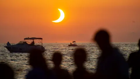 The eclipse shines in a crescent shape towards its end with silhouetted people on a beach in Ibiza