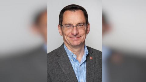 A man with short black hair in a suit. He is wearing glasses and has a baby blue chequered shirt under his blazer. 