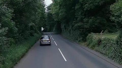 The road approaching Greeba Castle in Crosby. There is greenery on both sides of the road and an orange TT sign showing the bend in the area. There a a silver car on the road ahead.