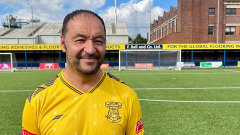 A man in a yellow football shirt is stood on a football pitch. The goal stands behind him, with a standing terrace behind that, and two factory units behind that. The man has short bark hair, dark stubble and his football shirt has the crest of Leek Town FC on the left chest.