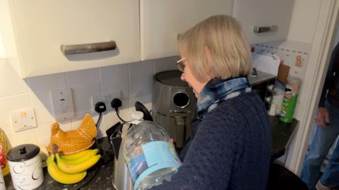 An elderly lady, wearing a scarf around her neck, stands in her kitchen pouring water from a large bottle into a kettle.