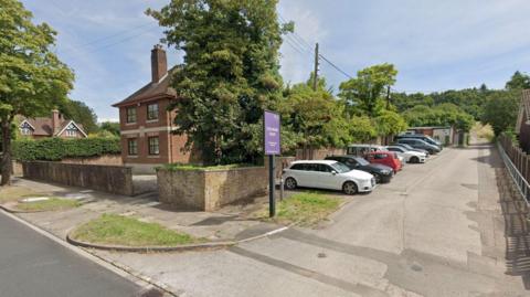 A Google street view image showing a two-storey house behind a tree and a wall. A purple sign saying "Collinson Court" is standing on a green patch next to the wall. A number of cars can be seen parked in a line by the wall.