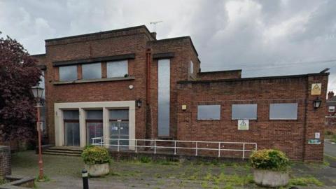 A flat roofed brick building with shutters at the windows and vegetation growing in between flag stones in front of the building.