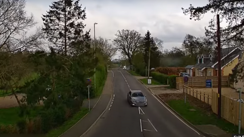 A silver car drives through a residential area with a police car behind it in pursuit 