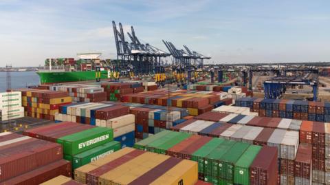 A view over the Port of Felixstowe. Large shipping containers can be seen stacked up next to each other and on top of each other. Container ships can be seen in the distance.