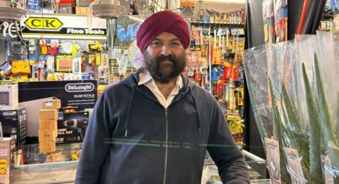 All Seasons DIY owner Surinder Josan stands in his shop looking at the camera, with items hanging from the ceiling and racks around the walls