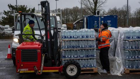 A man in a yellow high vis jacket driving a fork lift carrying stacks of bottled water. Another man in an orange high vis jacket is helping unload the water.