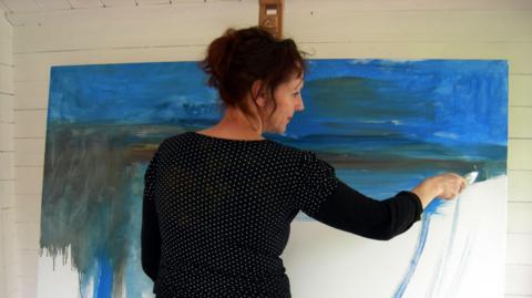 Woman with brown curly hair in a black top with small white dots seen from behind, with her face turned to the right, painting a canvas on an easel. She is painting an abstract, with blocky blue and green colours.