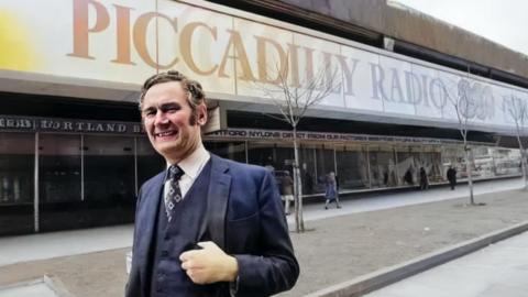 Philip Birch stands outside Piccadilly Radio's studios. He's wearing a suit including waistcoat. The Piccadilly Radio 261 text and logo adorns the outside window behind him.