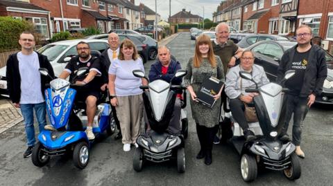 Nine people in the middle of a road. Residential houses are on either side of the road and cars are parking in front of the houses on the road and paths. From left to right: A man wearing a black zip-up hoodie, a light grey t-shirt, blue jeans and black trainers. He has glasses. A man sat on a blue mobility scooter wearing a black t-shirt and black shorts. He has glasses and short brown hair. A man with glasses stood wearing a white t-shirt, a yellow tie and a black jacket. A woman with shoulder-length brown hair and a fringe. She is wearing a white t-shirt, beige trousers and white shoes. A man sat on a black mobility scooter wearing a red t-shirt and a navy jacket with his arms crossed. A woman in a knee-length green and black spotty dress holding a Liberal Democrats clipboard. She has long ginger hair with a fringe. A man with grey hair stood wearing a green t-shirt. A woman sat on a black mobility scooter wearing an off-white jacket, black leggings and black trainers. A man with glasses wearing a black Liberal Democrats t-shirt, black jeans, beige shoes and a black zip-up jacket.   