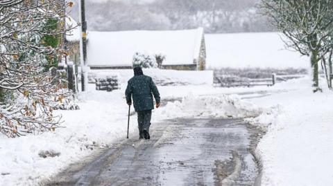 Man walks on a road partially cleared of snow, with a covering of snow on trees in the foreground and a barn and fields in the background