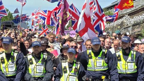 Police officers standing in front of a group of protesters who are holding a variety of flags, including the Union Jack and the flag of St George.