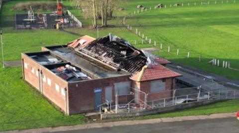 A picture showing the aftermath of the football club house following a fire. The roof is partly burned with the beamwork exposed. It is surrounded by green land and there is a children's play park in the background.