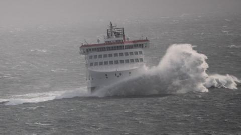 A large wave breaking against the bow of the Manxman ferry in rough seas. It is a large white red and black ferry and the sea surrounding it is grey with white-crested peaks.