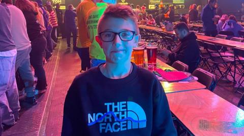 A boy with short hair and glasses, with a dark jumper which says The North Face, stands and smiles. Behind him are people standing up and sitting down at lines of tables in a hall.