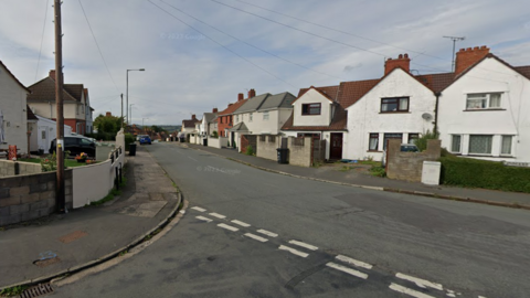 A screenshot from Google street view showing a junction on a residential road, with two-storey white houses and pavements on either side. 