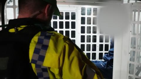 The back head and shoulders of a police officer is in the foreground on the left. In the background a suspect is sitting in a white barred enclosed cage in the back of a police van. His face is blurred.