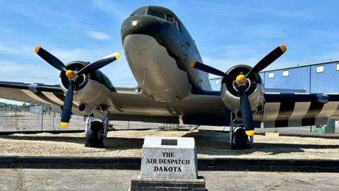 The restored Dakota Gate Guardian at RAF Brize Norton 47 Air Despatch Royal Logistic Corps in Oxfordshire on a sunny day. There is a marble badge and its name in front of the plane.