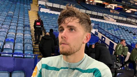 Mathias Kvistgaarden looks straight ahead and stands in front of a stand of blue seats. he wears a green zip up jacket.