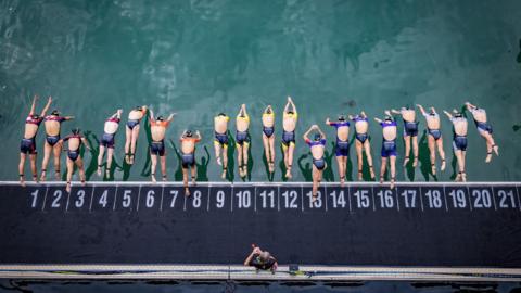 The image shows a group of swimmers lined up at the edge of a swimming pool, preparing to dive in. Each swimmer is positioned in a numbered lane (from 1 to 21), wearing swim caps and swimsuits. Some are mid-dive while others are poised to jump. An individual is standing on the pool deck.