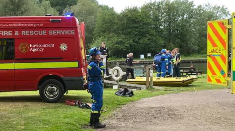 A red fire service van and an ambulance are parked by a river. A dinghy stands on the grass and a number of emergency service personnel are standing nearby.