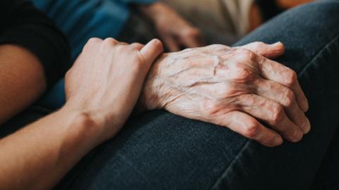 A young person's hand is resting reassuringly on an older persons hand.