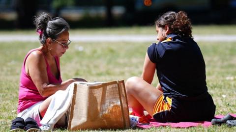 Two women sit on the grass with their shoes beside them and brown bag. 