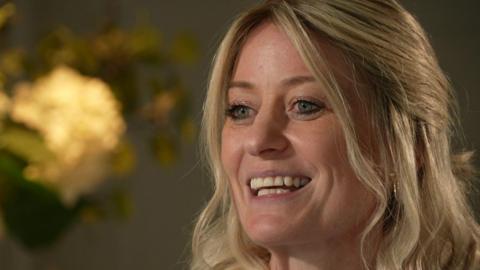 Charlotte Searle smiles as she's being interviewed for TV. She has shoulder-length blonde hair and there are yellow flowers in the backdrop on the left.