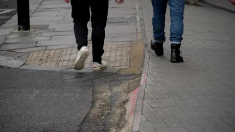 An uneven pavement with the feet of two people walking past. One wears white trainers and the other black boots.