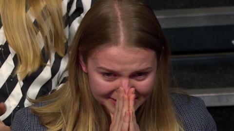 Woman with blond hair crying as she clasps her hands in front of her face