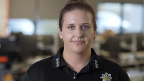Georgina Trownson smiles at the camera in a control room. She has dark hair that is tied up in a bun behind her head. She has hoop earrings and wears a black shirt. 