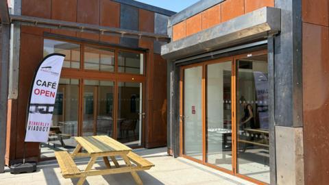 An orange steel building with glass doors and a flag which says Cafe Open
