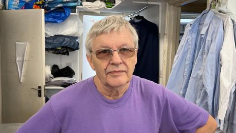 A picture of Rene stood in a launderette wearing a purple shirt. He has glasses on and has grey hair. He is stood in front of clothes that are hung up on the walls.