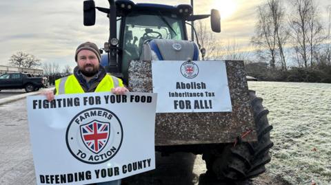 A man with brown hair and facial hair is wearing a brown jacket and a yellow hi-is jacket. He is holding a sign that reads "fighting for your food. Defending your county". Next to him us a blue tractor on a frosty path, there is a sign on it that reads " abolish inheritance tax for all"