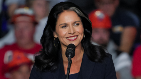 Former U.S. Rep. Tulsi Gabbard attends a campaign rally of Republican presidential nominee and former U.S. President Donald Trump at PPG Paints Arena in Pittsburgh, Pennsylvania - she is standing at the mic, has a broad smile, and gold earrings with a dark jacket and top. She has a distinctive white streak in her hair, and a blurred crowd can be seen behind her.