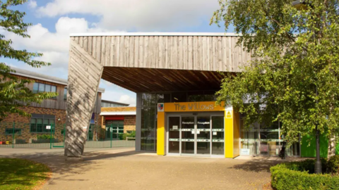 The school has a large wooden facade which spans the entrance and provides cover with a long piece of wood in keeping with the overall design touching the ground away from the entrance. The entrance is decorated yellow with glass doors and windows.
