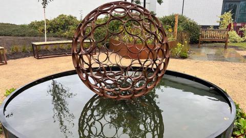 A scaled-down version of the circular garden, with a reflection pond in the centre with aspherical steel sculpture in the middle made up of rusted circles.  A gravel path surrounds it and there are benches in the background