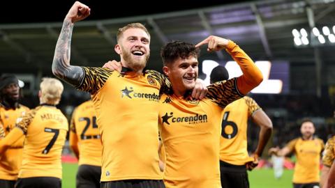 Hull City's Oliver McBurnie celebrates with Ryan Giles after scoring a goal at the MKM Stadium. McBurnie has blond hair and beard and Giles has black hair. The kit is amber shirts with black arm patterns and black shorts. Other Hull City players are in the background