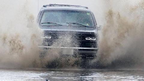 The picture shows a dark-coloured car driving straight through deep surface water, sending large walls of spray up on both sides of the vehicle. The water is being forced aside by the front of the car, creating a dramatic splash that almost reaches the height of the bonnet and partially obscures the headlights. The road surface is completely covered by water, suggesting local flooding rather than a small puddle.