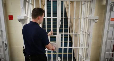 Prison officer wearing a dark uniform is about to lock a metal gate leading to some stairs in a jail.
