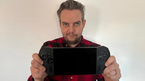Tom Gerken holding a ROG Xbox Ally X. It is a black handheld console, which looks like an Xbox controller cut in half with a screen in the middle.